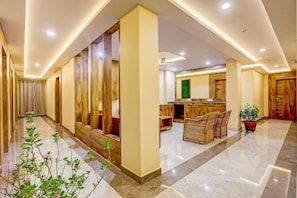 A hotel lobby with a marble floor, wooden paneling, a reception desk, wicker furniture, and a potted plant.