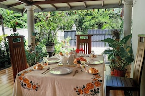 Uma área de refeições ao ar livre com uma mesa posta para dois, cercada por plantas em vasos e uma pérgola de madeira.