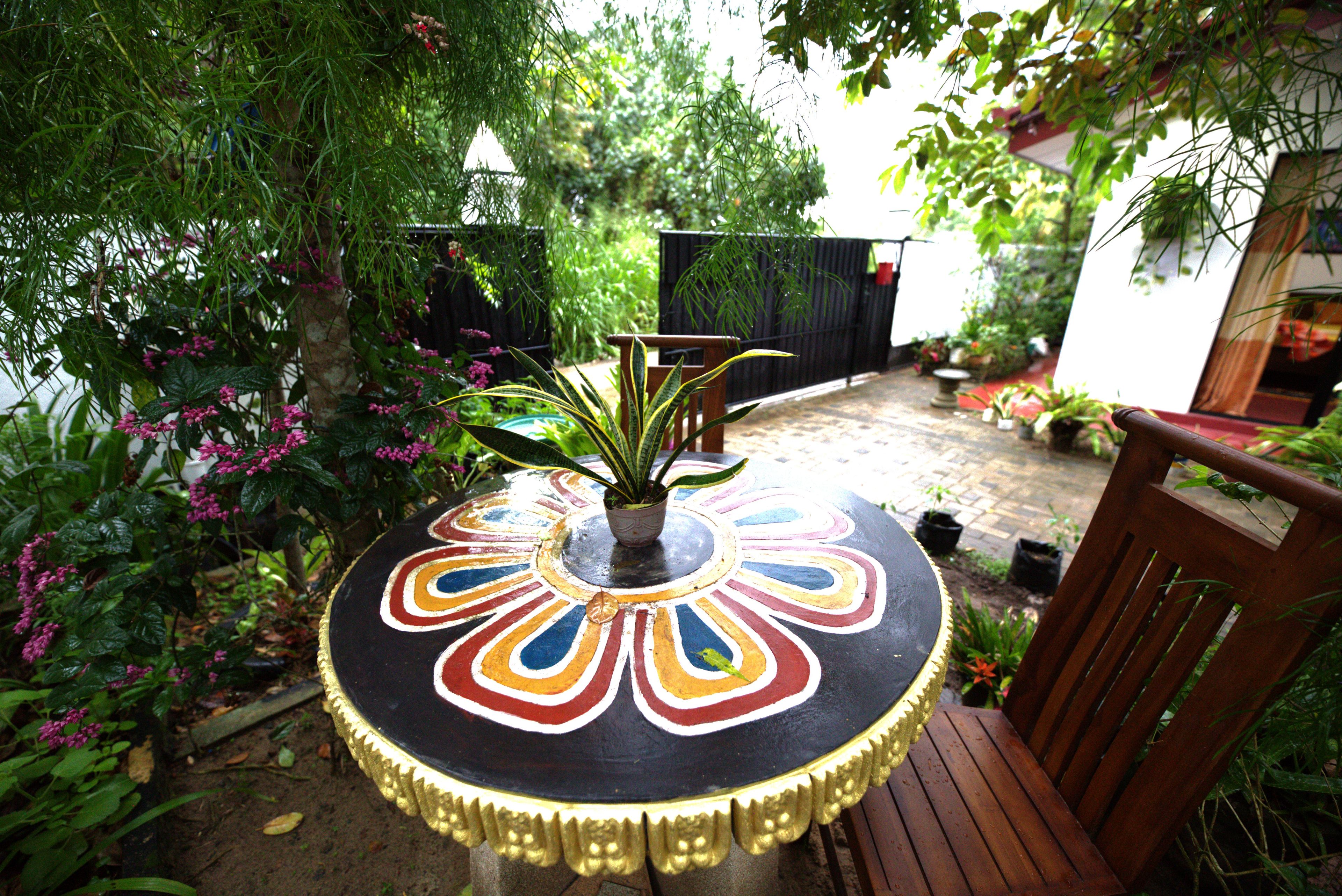 A round table with a colorful painted top, surrounded by a wooden chair, in a garden with various plants and a fence in the background.