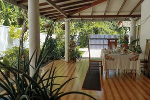 Uma área externa coberta com piso de madeira, uma mesa posta para refeições e diversos vasos com plantas.