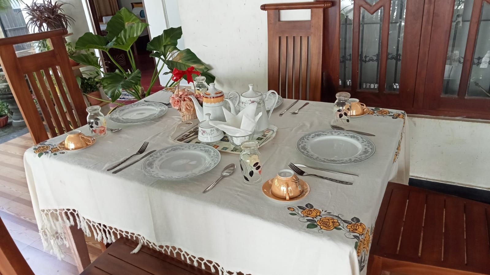 Une table à manger dressée avec des assiettes, des couverts et une théière, entourée de chaises et de plantes en pot.