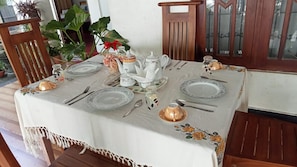 A dining table set with plates, cutlery, and a teapot, surrounded by chairs and potted plants.