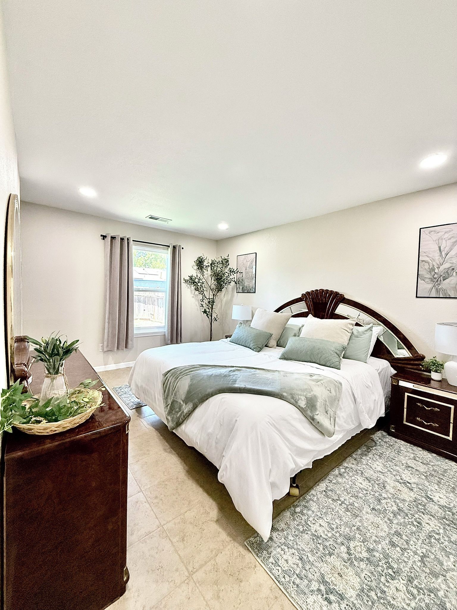A bedroom with a large bed, a bedside table, a nightstand, a window with curtains, a potted plant, and a wall-mounted artwork.