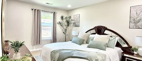 Un dormitorio con una cama grande, una mesita de noche, una lámpara, una ventana con cortinas, una planta en maceta y una obra de arte colgada en la pared.