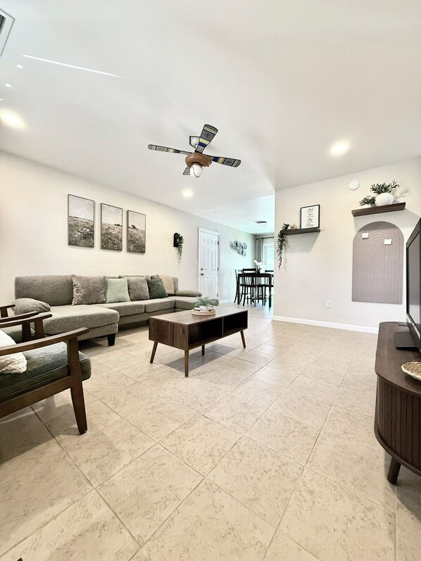 A spacious living room with a sofa, coffee table, and TV stand. There is a ceiling fan and a dining area in the background.