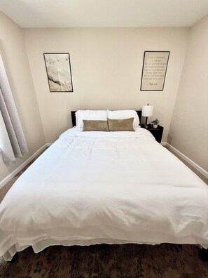 A neatly made bed with white linens and two pillows, a bedside table with a lamp and a small plant, and two framed quotes on the wall.