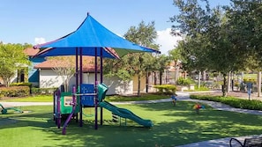 A playground with a slide and climbing structures, surrounded by greenery and a building in the background. - Bella Vida Home w Pool - Near All Parks (Kissimmee)