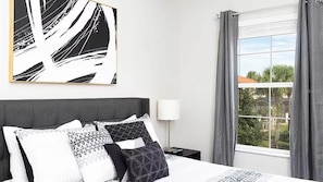 A hotel room with a bed, a bedside lamp, a window with curtains, and a wall-mounted abstract painting. - Bella Vida Home w Pool - Near All Parks (Kissimmee)
