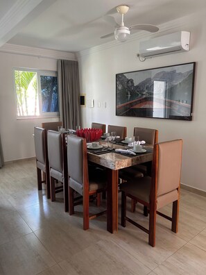 Uma sala de jantar com uma mesa de tampo em mármore, seis cadeiras, um ventilador de teto e uma grande moldura na parede.