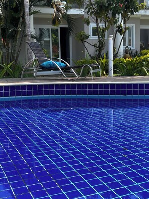 A swimming pool with a tiled edge, a lounge chair with a cushion, and a building with a door in the background. - CJ17AR/3D-Paraíso em Arraial D’ajuda/Praia e Lazer (Arraial da Ajuda)
