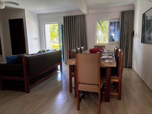 A dining area with a table set for four, a sofa, and a view of greenery outside. - CJ17AR/3D-Paraíso em Arraial D’ajuda/Praia e Lazer (Arraial da Ajuda)