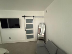 Ein Zimmer mit einem wandmontierten Flachbildfernseher, einer Schiebetür mit satinierten Glasscheiben, einem Bett mit weißem Kopfteil und einem grauen Sofa.