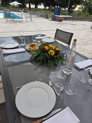 A table set for dining outdoors with a pool view, including plates, cutlery, glasses, and a bottle.