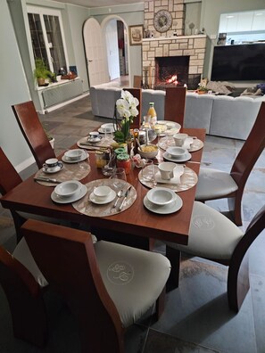 A dining area with a wooden table set for four, surrounded by chairs with embroidered logos. A fireplace is visible in the background, and a television is mounted on the wall.