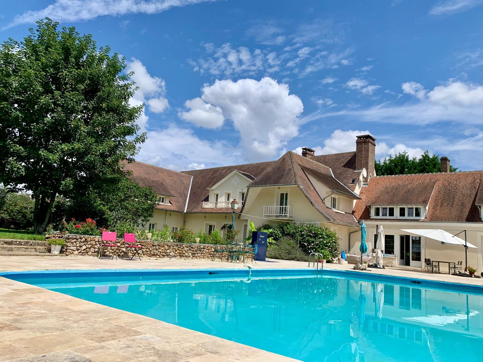 A swimming pool with a stone deck, surrounded by a garden and a large house with a pitched roof.