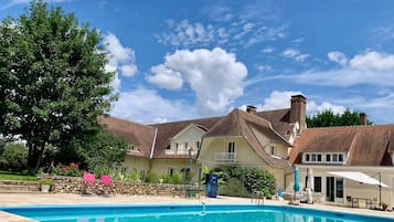 A swimming pool with a stone deck, surrounded by a garden and a large house with a pitched roof.