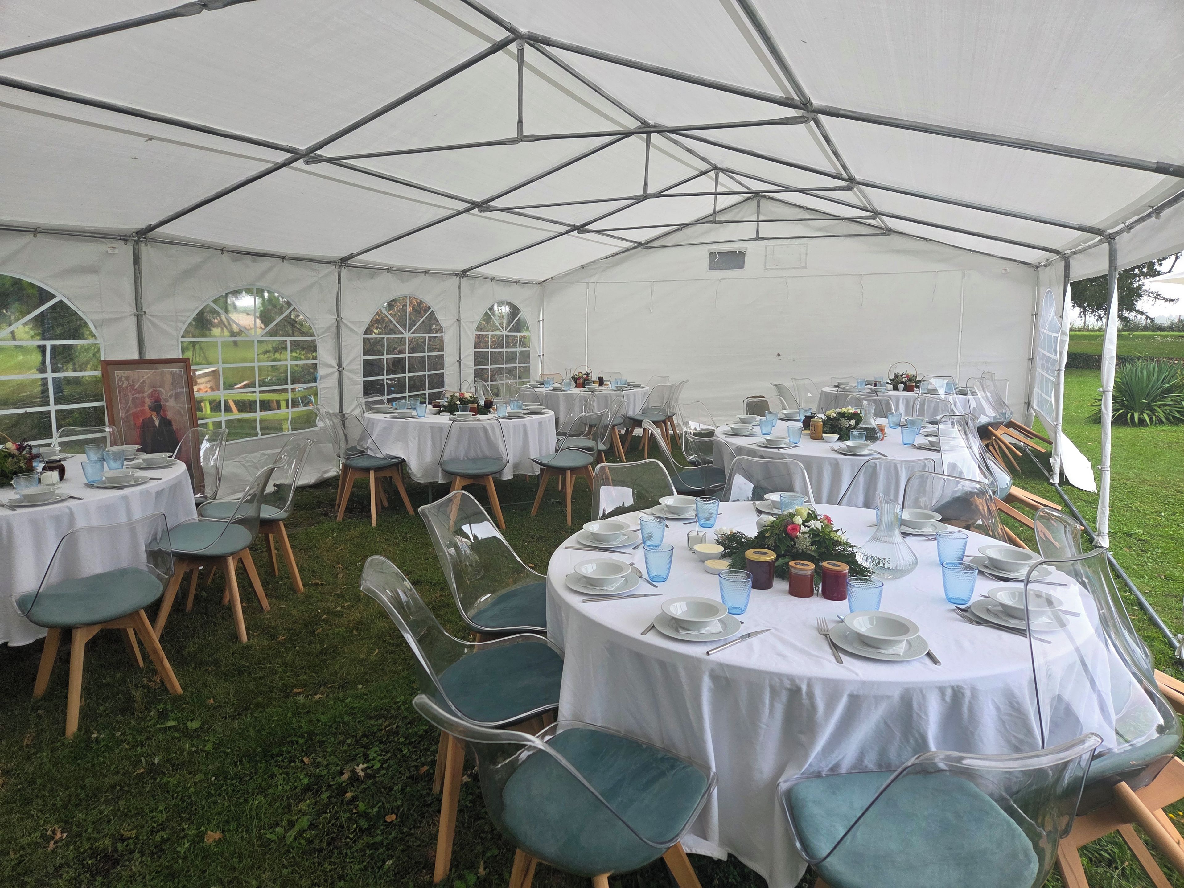 Un espace de restauration en plein air, aménagé sous une tente, avec des tables rondes, des chaises transparentes et des nappes blanches.