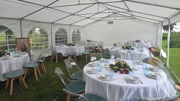 Un espace de restauration en plein air, aménagé sous une tente, avec des tables rondes, des chaises transparentes et des nappes blanches.