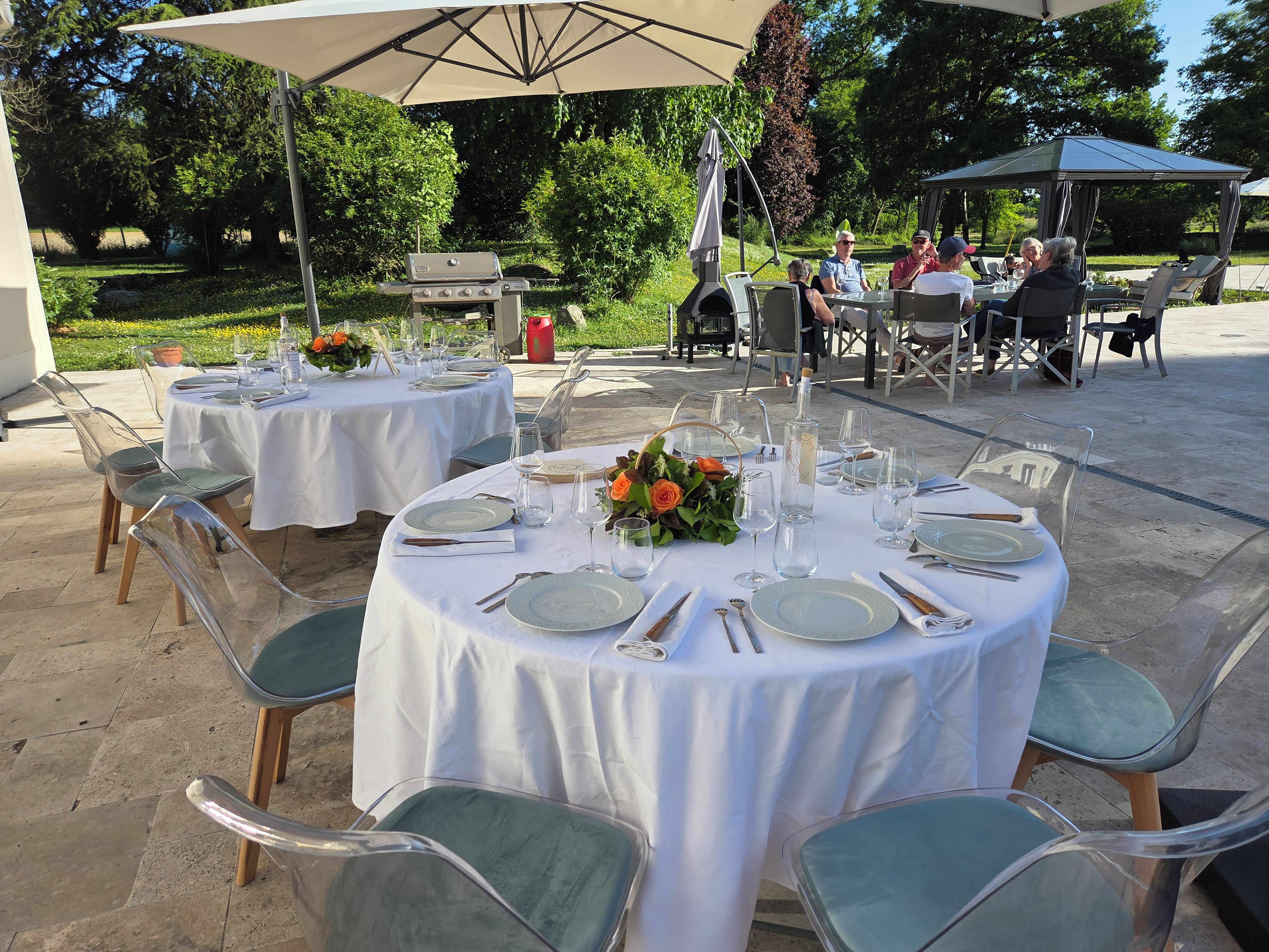 Outdoor tables set up with white tablecloths, clear chairs, and a barbecue grill.