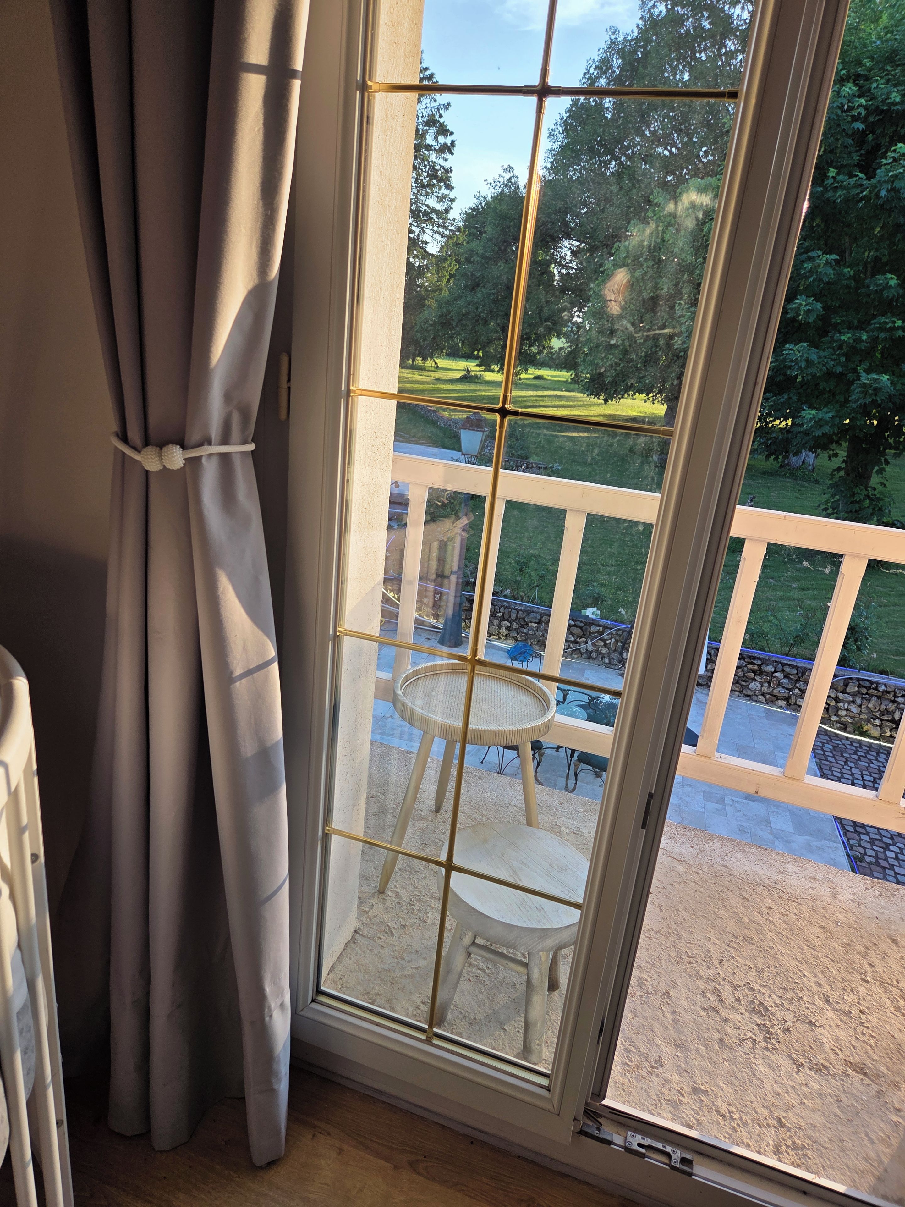 Ein Zimmer mit großem Fenster, Vorhängen und Blick auf eine Terrasse mit Stuhl und Tisch.