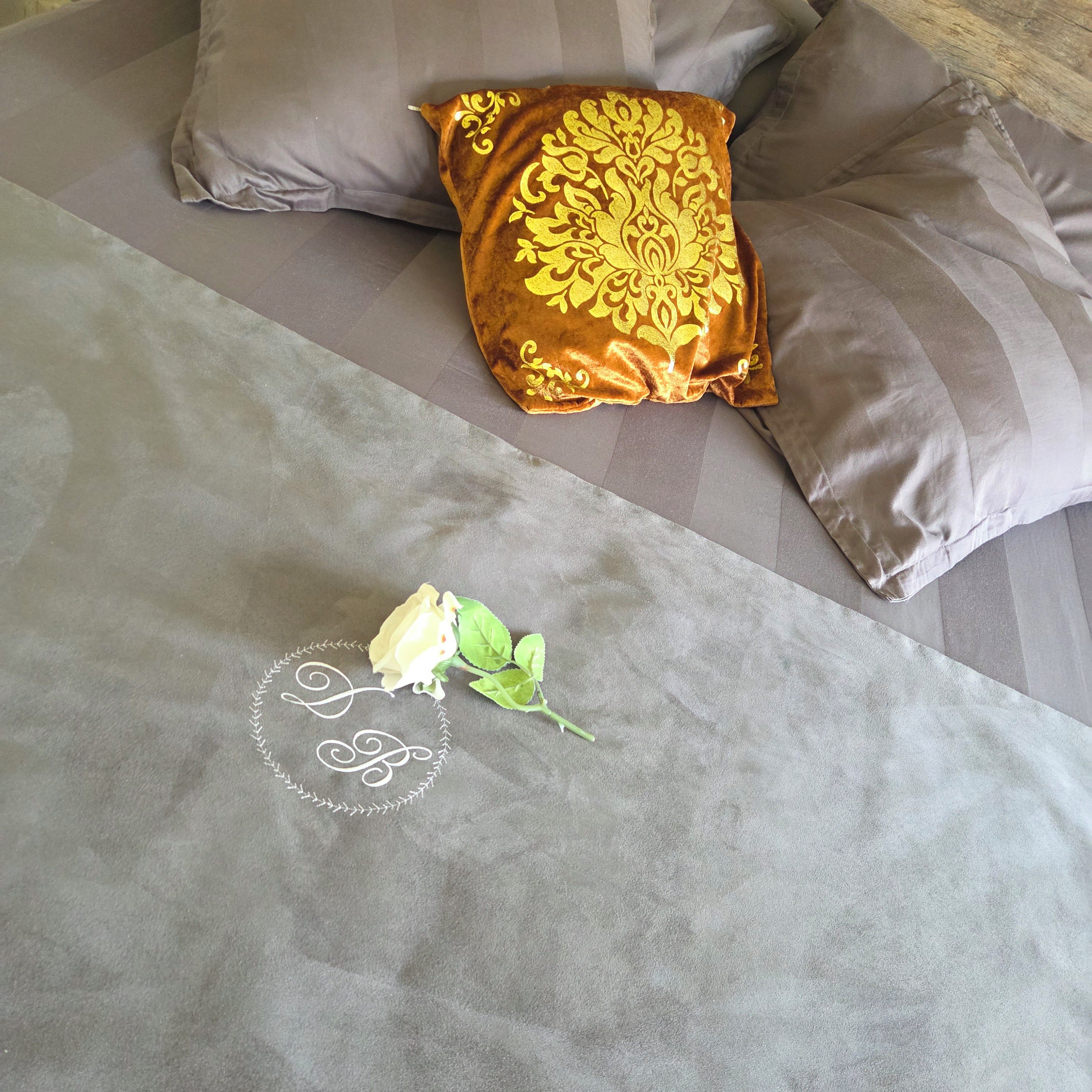 Uma cama com colcha cinza, um travesseiro decorativo com padrão dourado e uma única rosa branca.