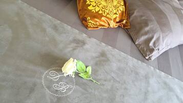 Uma cama com colcha cinza, um travesseiro decorativo com padrão dourado e uma única rosa branca.