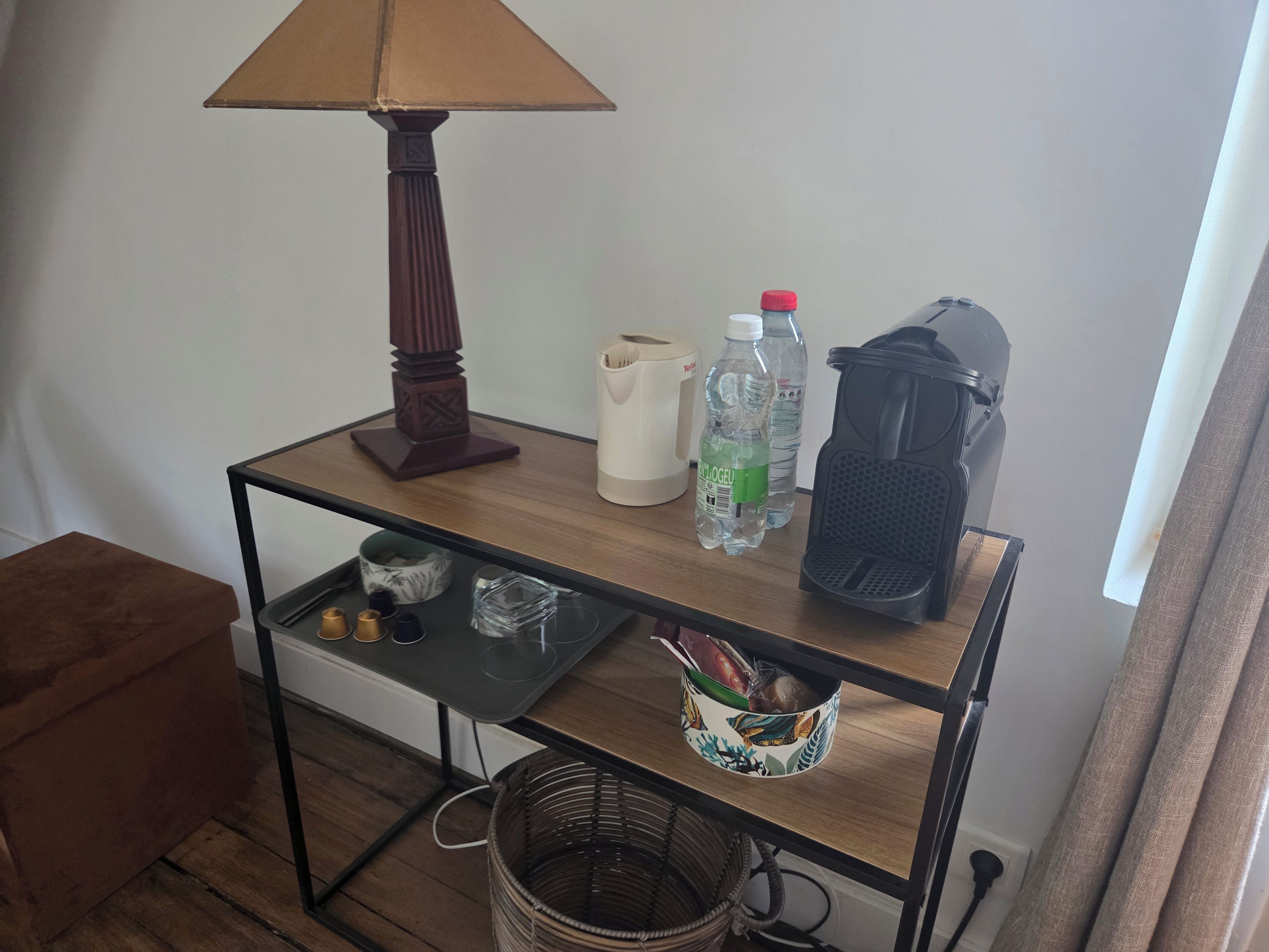 A wooden table with a lamp, water bottles, a kettle, and a basket with snacks.