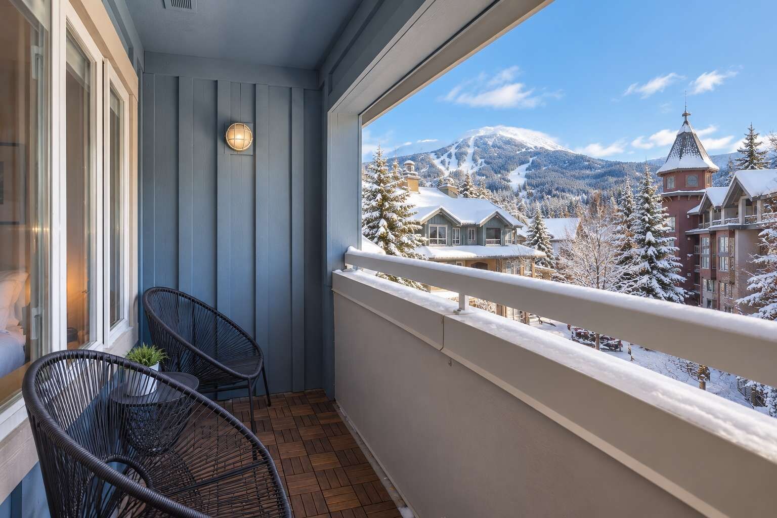 A balcony with two wicker chairs and a small table, overlooking a snowy mountain landscape with buildings.