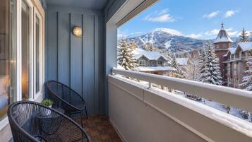 A balcony with two wicker chairs and a small table, overlooking a snowy mountain landscape with buildings.