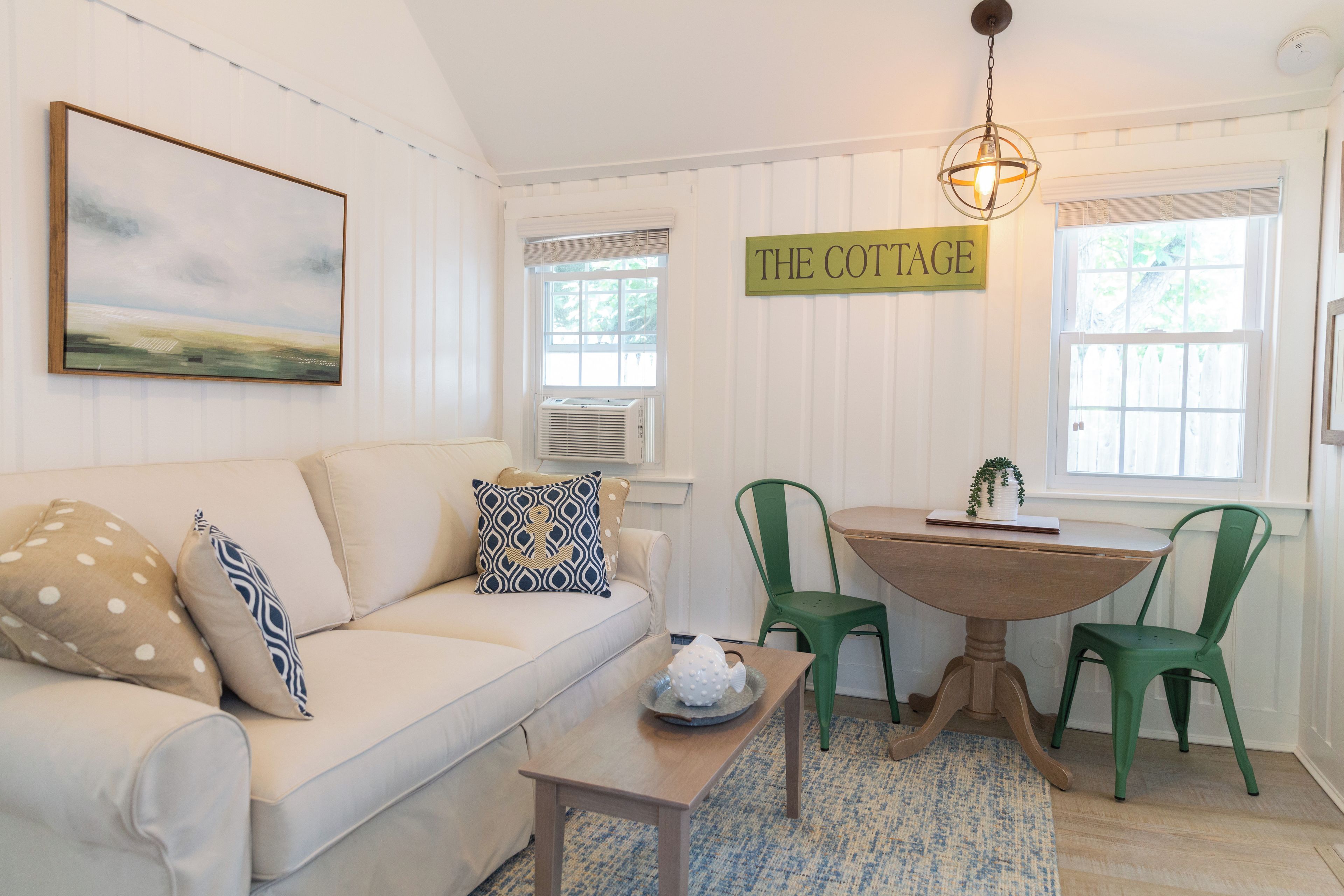 A cozy living room with a white sofa, a coffee table, and a dining table with chairs. A painting and a sign reading 'THE COTTAGE' are on the wall.