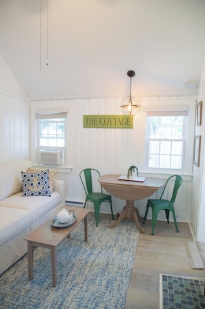 Ein gemütliches Cottage-Interieur mit einem weißen Sofa, einem Holztisch, grünen Stühlen und einem Schild mit der Aufschrift 'THE COTTAGE' an der Wand.