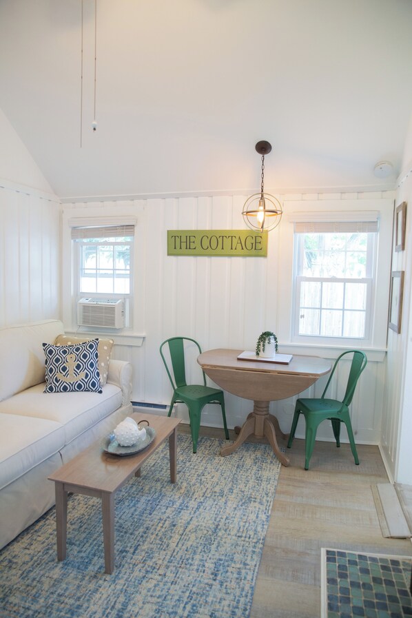 Interior acogedor de una cabaña con un sofá blanco, una mesa de madera, sillas verdes y un letrero en la pared que dice 'THE COTTAGE'.