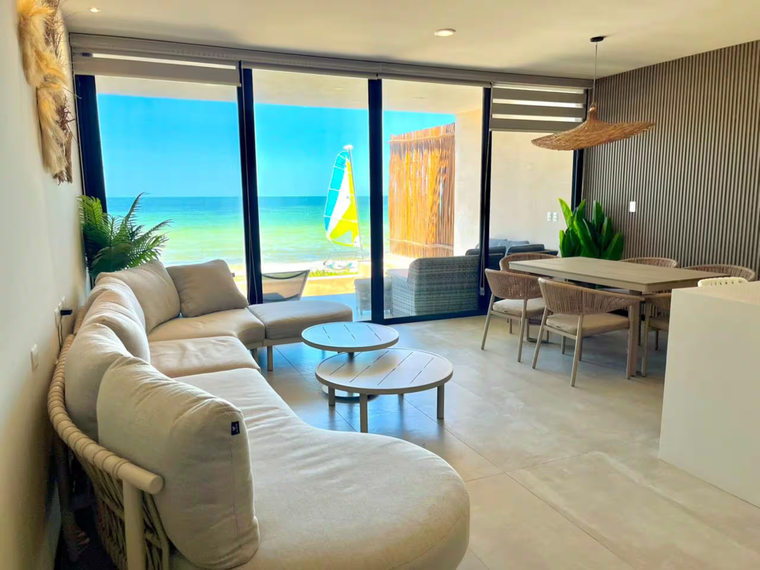 A modern living room with a sofa, coffee table, and dining area, overlooking a beach with a sailboat.