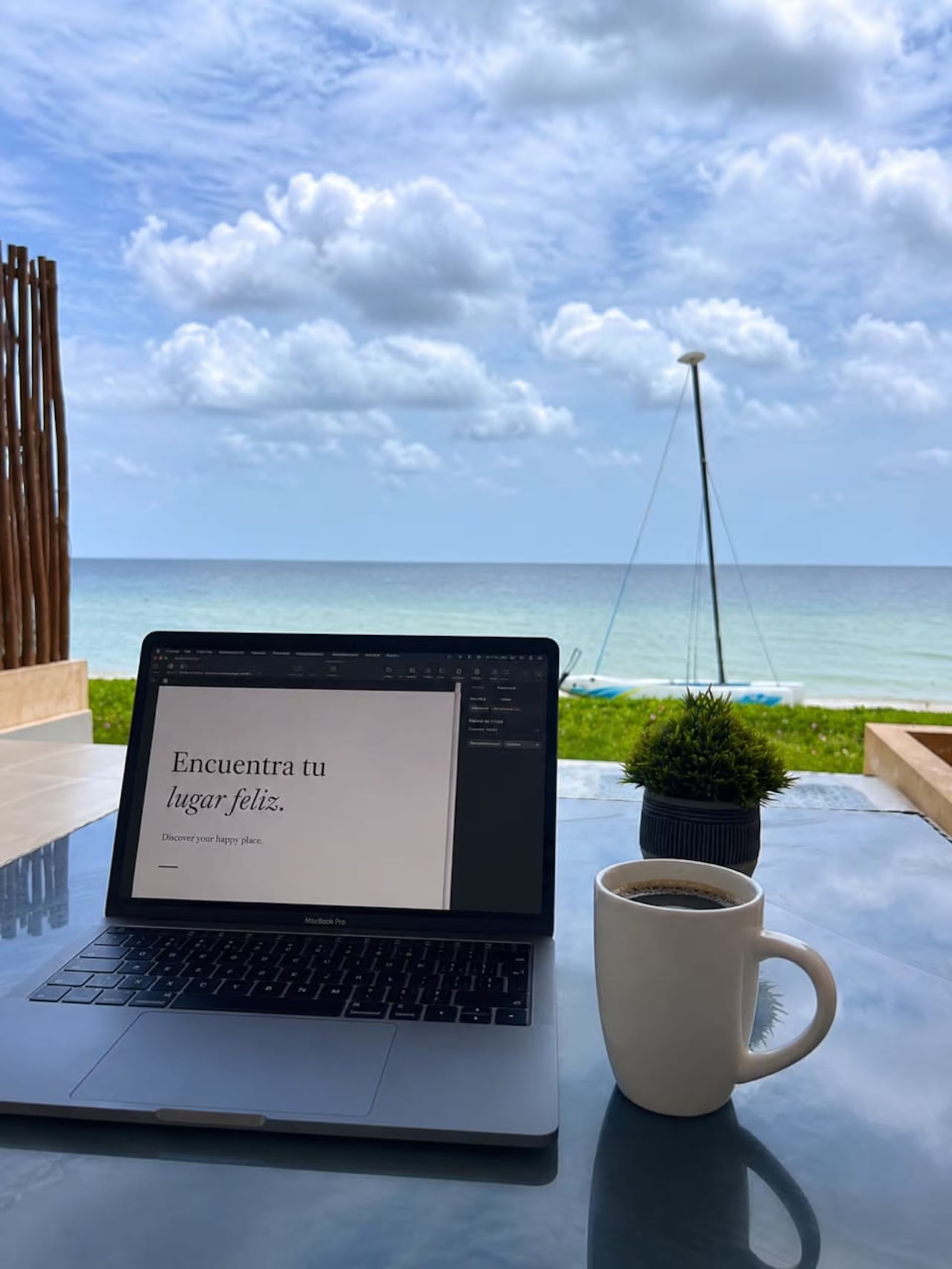 A laptop on a table with a message on the screen, a cup of coffee, and a view of the ocean and a sailboat in the background.