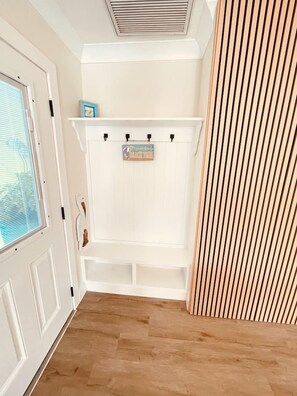 Habitación de esquina con una puerta blanca, una ventana con persianas y un estante blanco fijado a la pared con un letrero, además de suelo de madera.