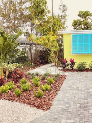 A landscaped garden with a paved walkway leading to a building with blue shutters. - Waterfront Bungalow Getaway | Sleeps 12 (Madeira Beach)