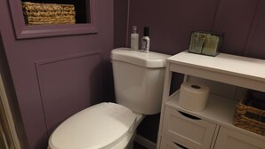 A bathroom with a white toilet, a white vanity with drawers, a basket on the wall, and a roll of toilet paper on the vanity. - Knot Northern Retreat & Spa: Hot Tub • Gym • Workspace (Minneapolis)