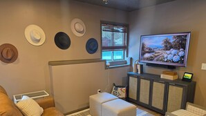 A living room with a brown leather sofa, a television on a stand, a cabinet with decorative items, and a window with blinds. - Knot Northern Retreat & Spa: Hot Tub • Gym • Workspace (Minneapolis)
