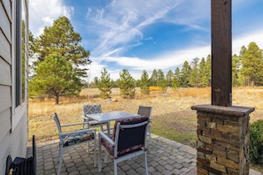An outdoor patio with a table and chairs, a stone pillar, and a view of a grassy field with trees. - Wolfmoor spacious cabin with trail access (Flagstaff)