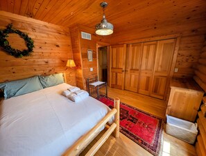 A wooden cabin bedroom with a bed, a nightstand, a lamp, a wardrobe, and a red patterned rug. - Skyline Log Cabin • River Views + Screened Patio (Wabasha)