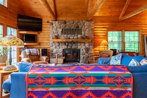 A log cabin interior with a stone fireplace, wooden walls, a television mounted on the wall, and a colorful blanket on the couch. - Skyline Log Cabin • River Views + Screened Patio (Wabasha)