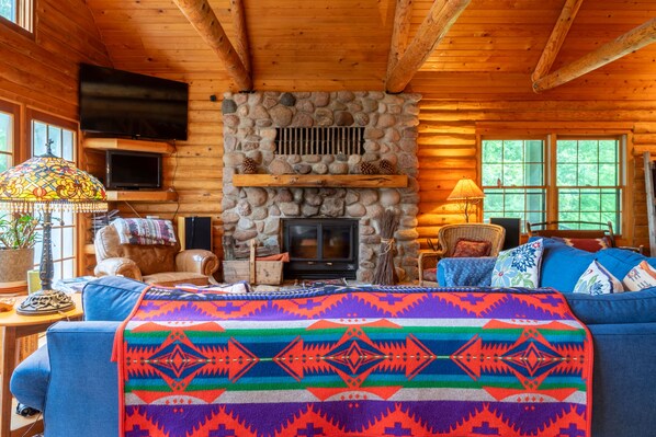A log cabin interior with a stone fireplace, wooden walls, a television mounted on the wall, and a colorful blanket on the couch.