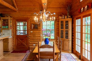 A wooden interior with a dining table, chairs, a chandelier, and large windows.