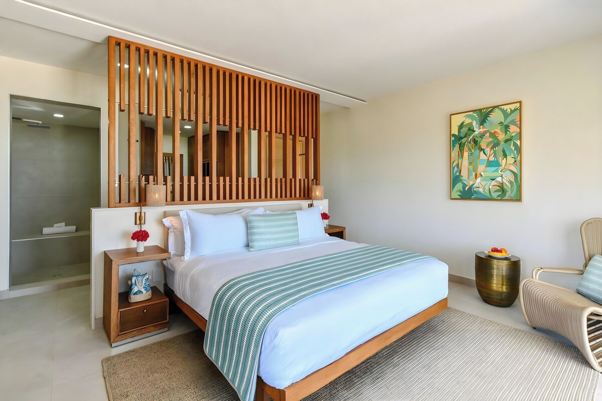 Un dormitorio moderno con una cama grande, cabecera de madera, mesitas de noche, un tocador, una silla, un cuadro y un baño con cabina de ducha de vidrio.