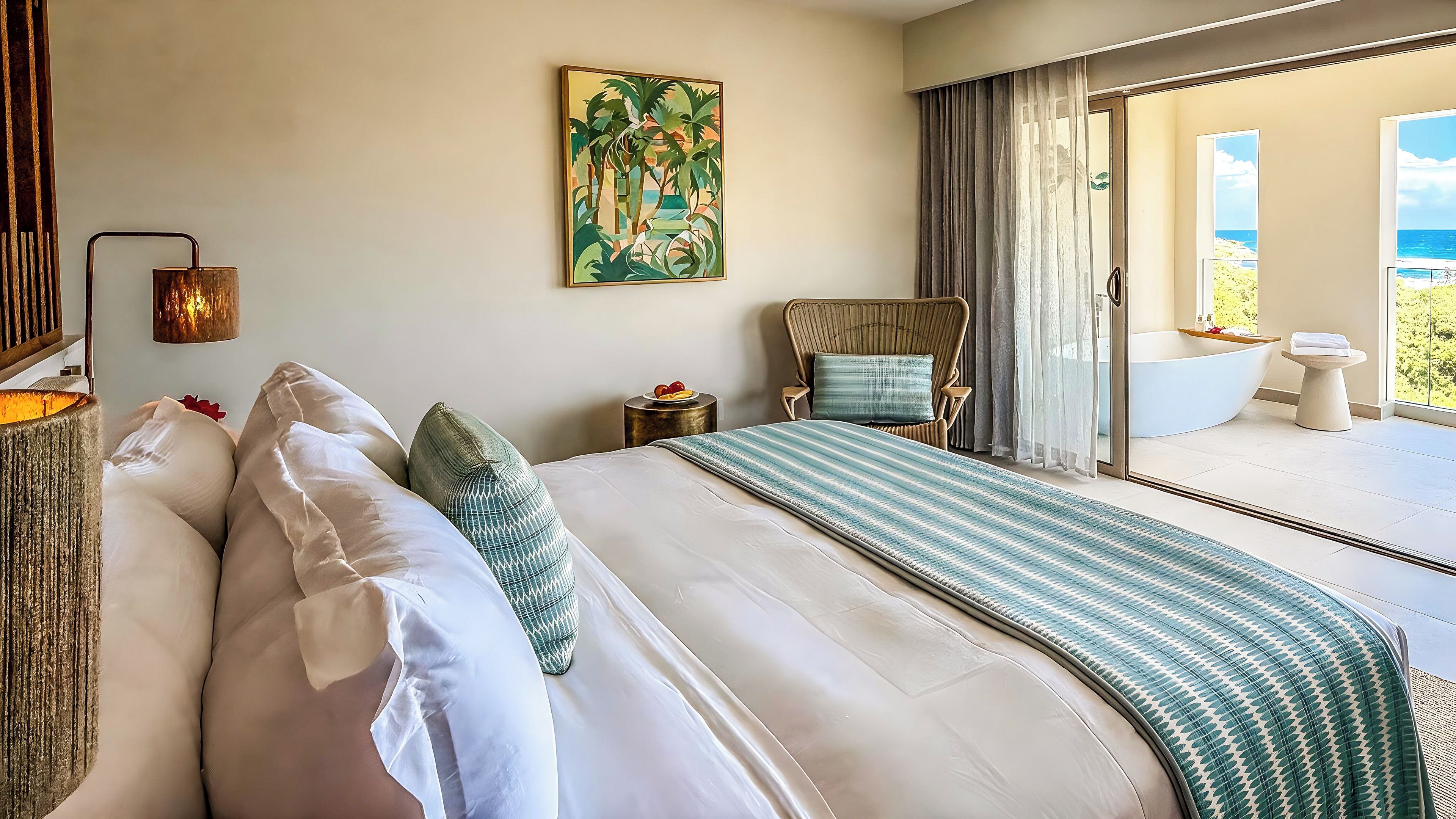 A hotel room with a large bed, a view of the sea, a painting on the wall, and a bathroom with a bathtub.