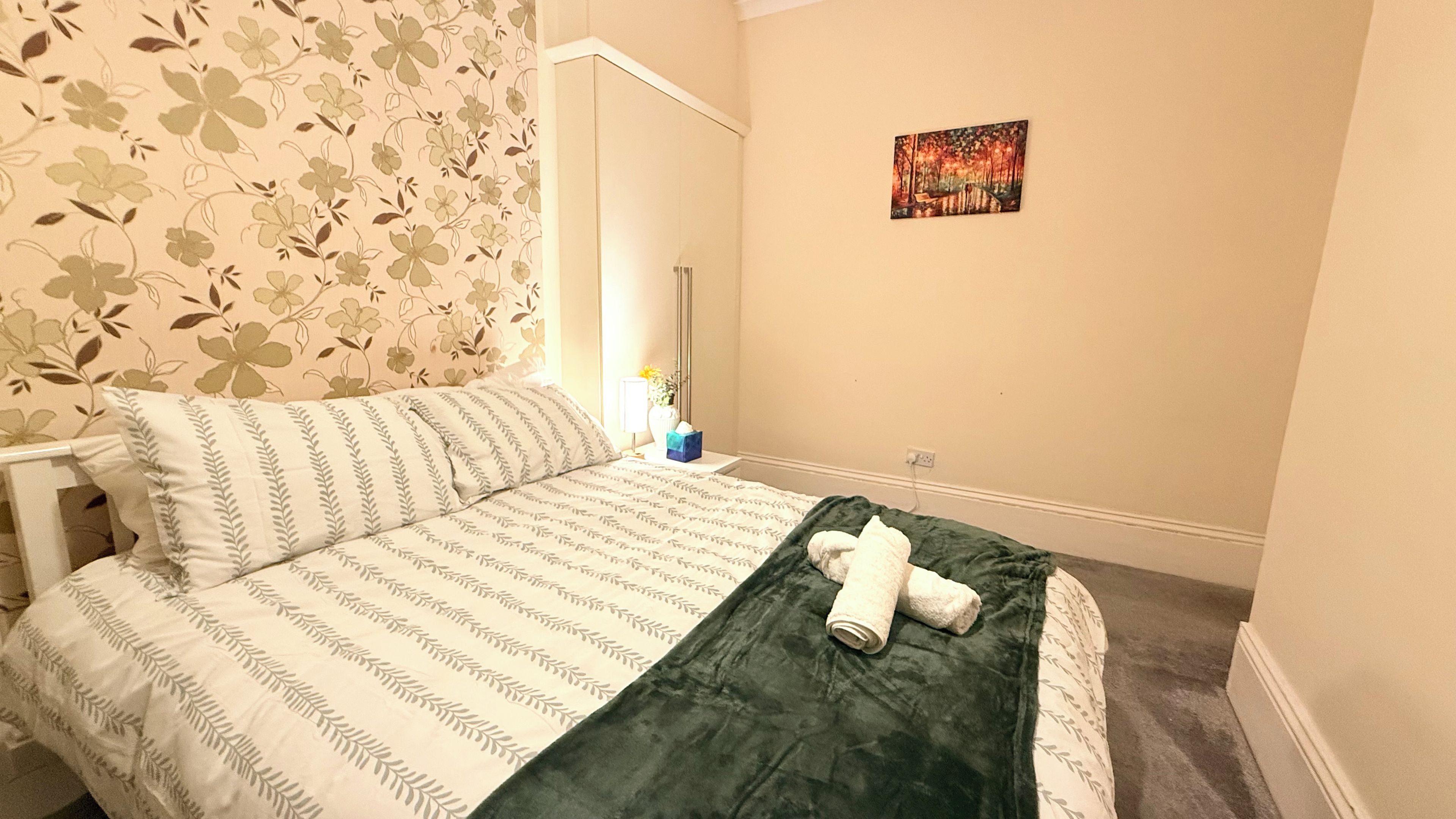 A hotel room with a bed, a green blanket, a painting on the wall, and a window with curtains.