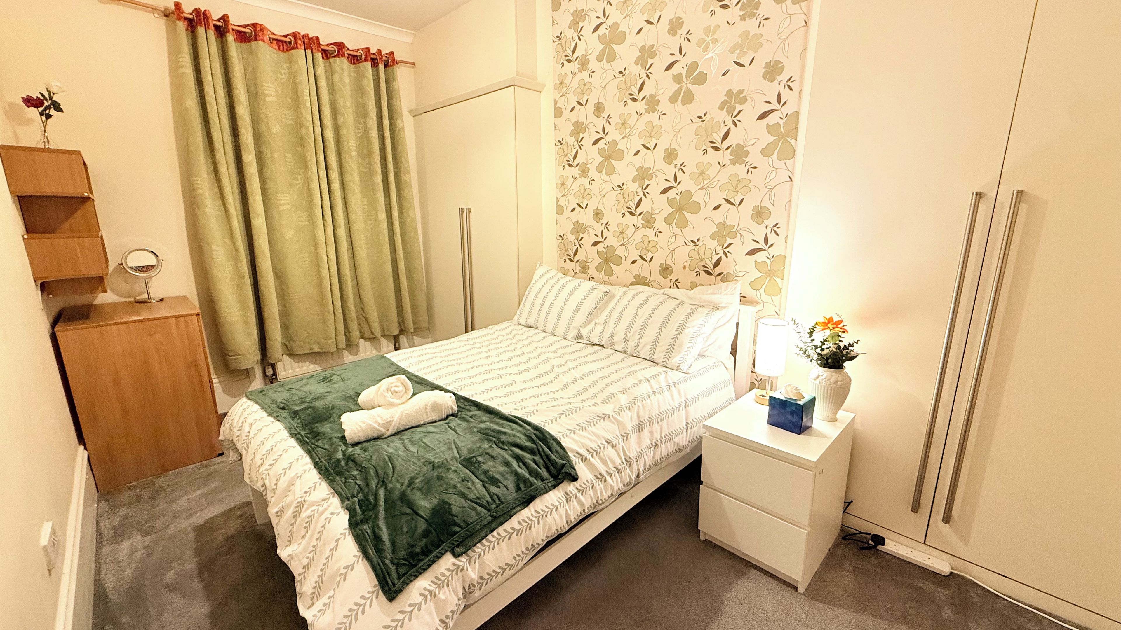 A neatly made bed with a green blanket and a white towel, a bedside table with a lamp and a vase of flowers, and a wardrobe with a floral wallpaper.
