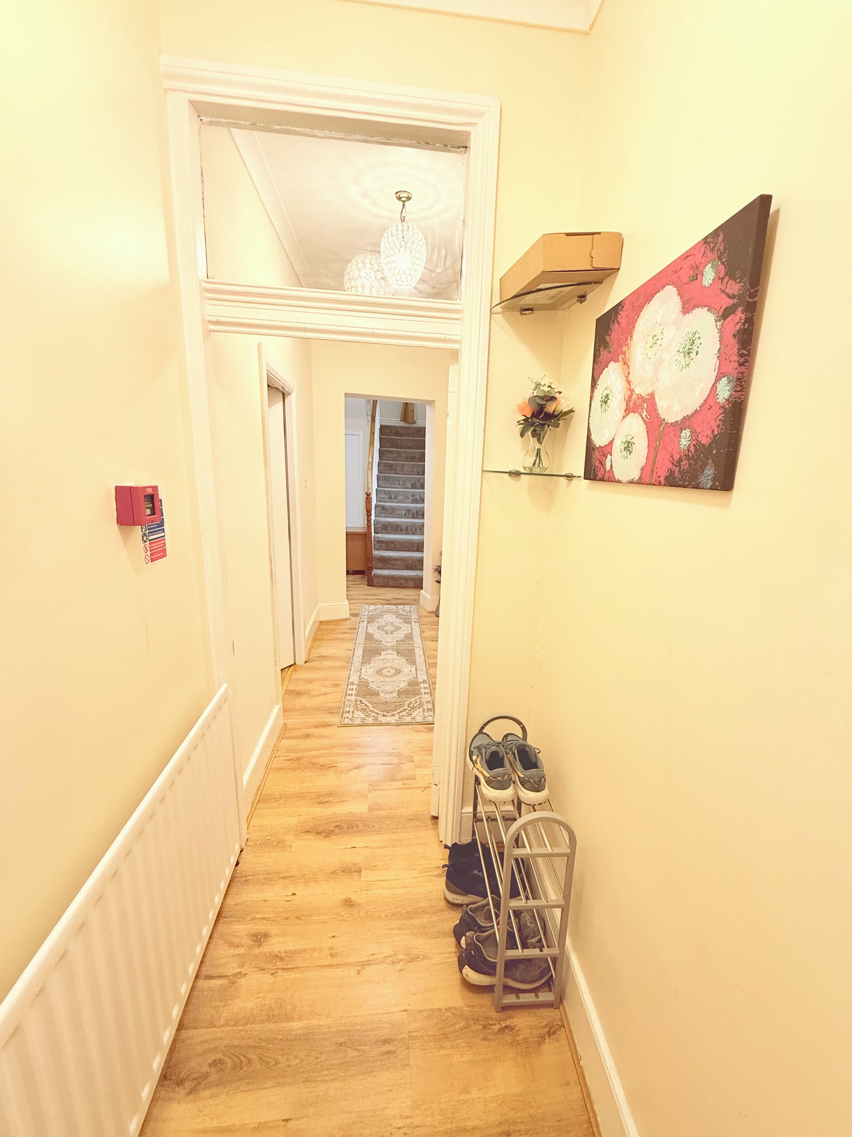A hallway with a wooden floor, a shoe rack, a painting, and a staircase in the background.