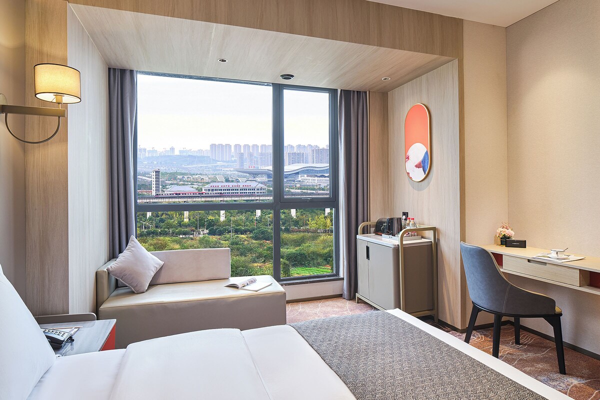 A hotel room with a large window, a bed, a desk, a chair, and a small cabinet.