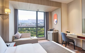 A hotel room with a large window, a bed, a desk, a chair, and a small cabinet.
